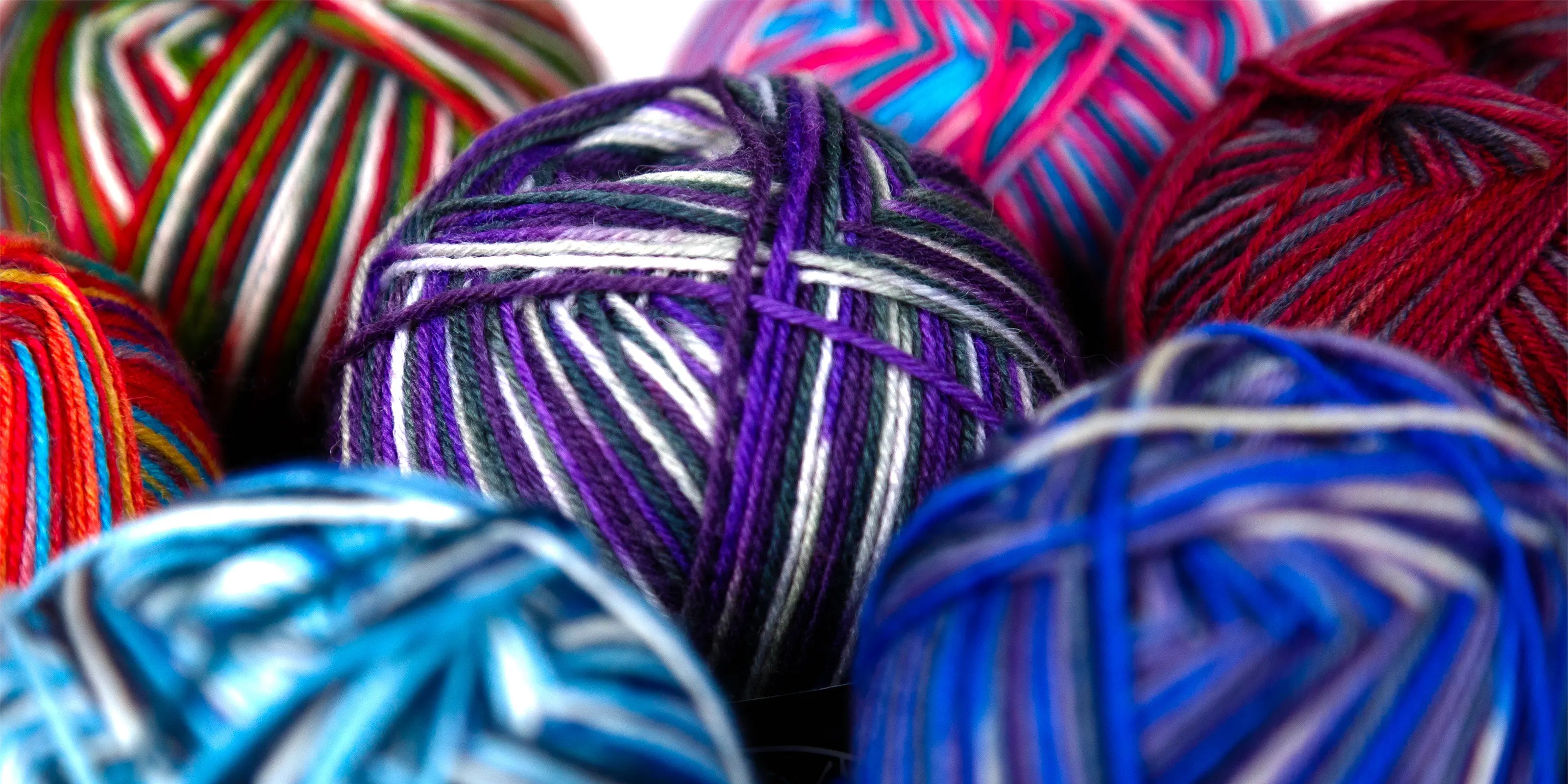 Angled depth-of-field photo showcasing Sock Flock yarn balls including Sea Glass, Purple Ivy and Blue Moon, with bold stripe blends and soft texture detailing.