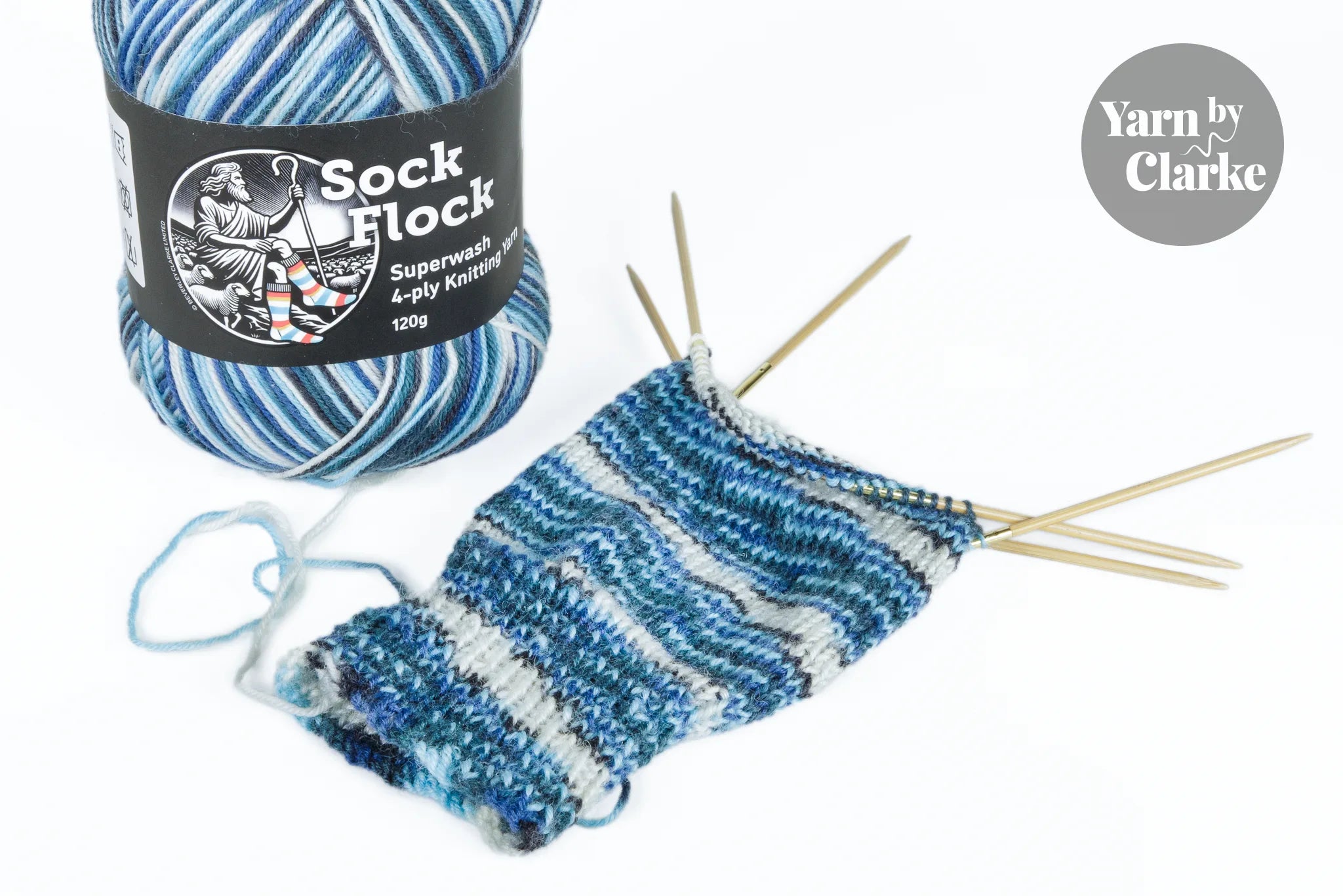 A half-knitted blue sock on double-pointed needles beside a ball of Sock Flock yarn, illustrating pattern development during knitting.